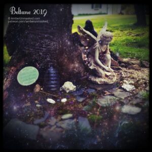 In front of the stump is the largest fairy who is new to the group, Fiona Brightwing. On the ground around her, there are slate stones, mossy patches, jewels, a decorative glass bottle, and a small sign. Photos taken on Beltane (May 1, 2019). Glittery sparkles are all around the area.