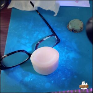 desk covered in a blue silicone working mat; on top out of focus is the aqua colored aluminum lid of the jar discovered; a pair of reading glasses; the Pond's milk glass jar upside-down; a black light illuminating the jar and also showing that the blue mat is covered in invisible substances (epoxy resin).