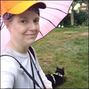 Amber wearing white shirt, neon orange baseball hat, holding a pink parasol white black cat Gus lies in the grass.