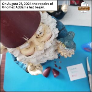 garden gnome with blue shirt, green pants, red conical hat, holding a bluebird; Gnomez Addams on top of a desk to be repaired. "On August 27, 2024 the repairs of Gnomez Addams hat began."