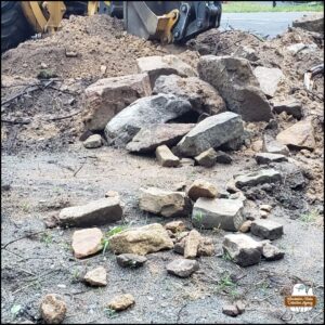 the excavation of the end of the upper parking lot with the beginning stages of building a new rock wall; the excavator machine mostly out of frame next to a pile of dirt and rocks.