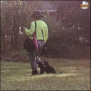 caught on trailcam in the daylight, black cat Gus sitting in the grass as biographer Amber stands next to him holding her camera, camera bag, evidence baggies, cell phone, and pink parasol.