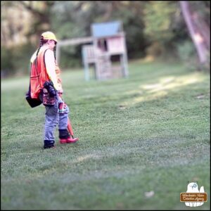 Amber wearing exploring clothes (jeans, boots, bright neon yellow shirt, safety orange vest and cap, with bags and camera) on trailcam.