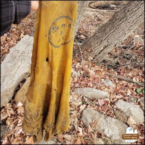 held up by Amber's hand at the edge of the woods: a piece of yellow fabric with lettering: V.F.M.A & J.C. inside a circle. The fabric is filthy and has holes. It was buried between rocks that had been excavated and moved. It looks more like it could have been a laundry bag rather than a shirt.