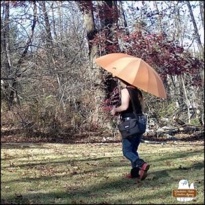 Amber wearing jeans, red boots, a black tank top, a camera bag hanging from left shoulder, face mostly hidden by the large orange umbrella.