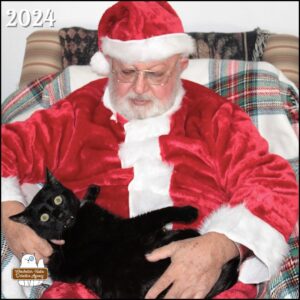 black cat Gus with his fangs showing and huge (alarmed) eyes while Santa holds him like a baby.