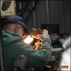 Over the right shoulder of the Grumpy Old Man at a workbench sharpening a chainsaw chain.